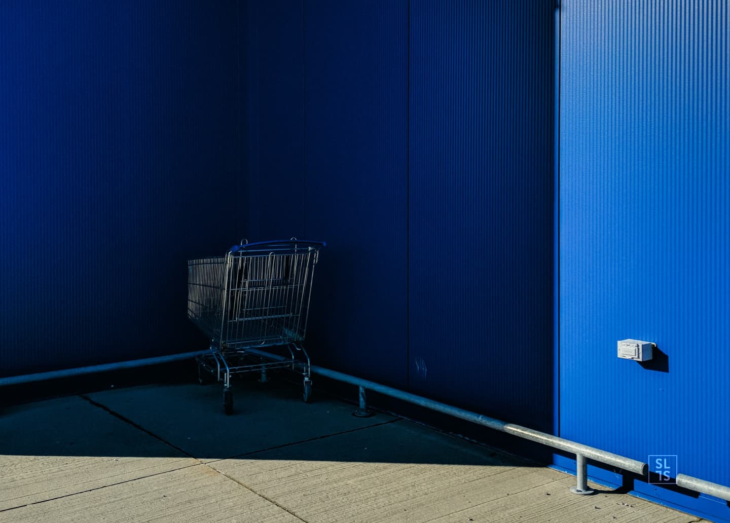 Chariot de course blotti dans l'ombre, devant une façade bleue, symbole du panier abandonné pendant le parcours sur un site e-commerce