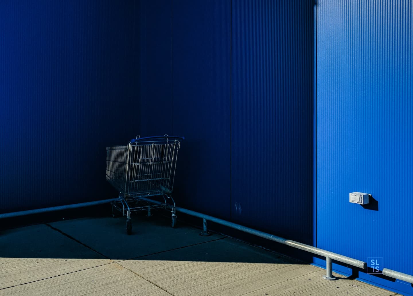 Chariot de course blotti dans l'ombre, devant une façade bleue, symbole du panier abandonné pendant le parcours sur un site e-commerce